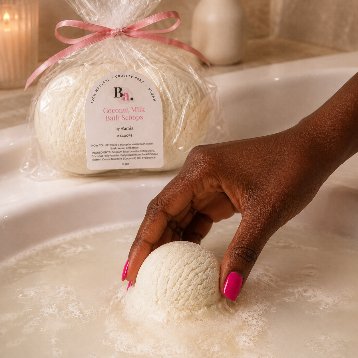 A hand with bright pink nails holds a round cream-colored Coconut Milk Bath Scoop over foaming warm water in a white tub, with a packaged bag of by A'amia Coconut Milk Bath Scoops tied with a pink ribbon in the background.