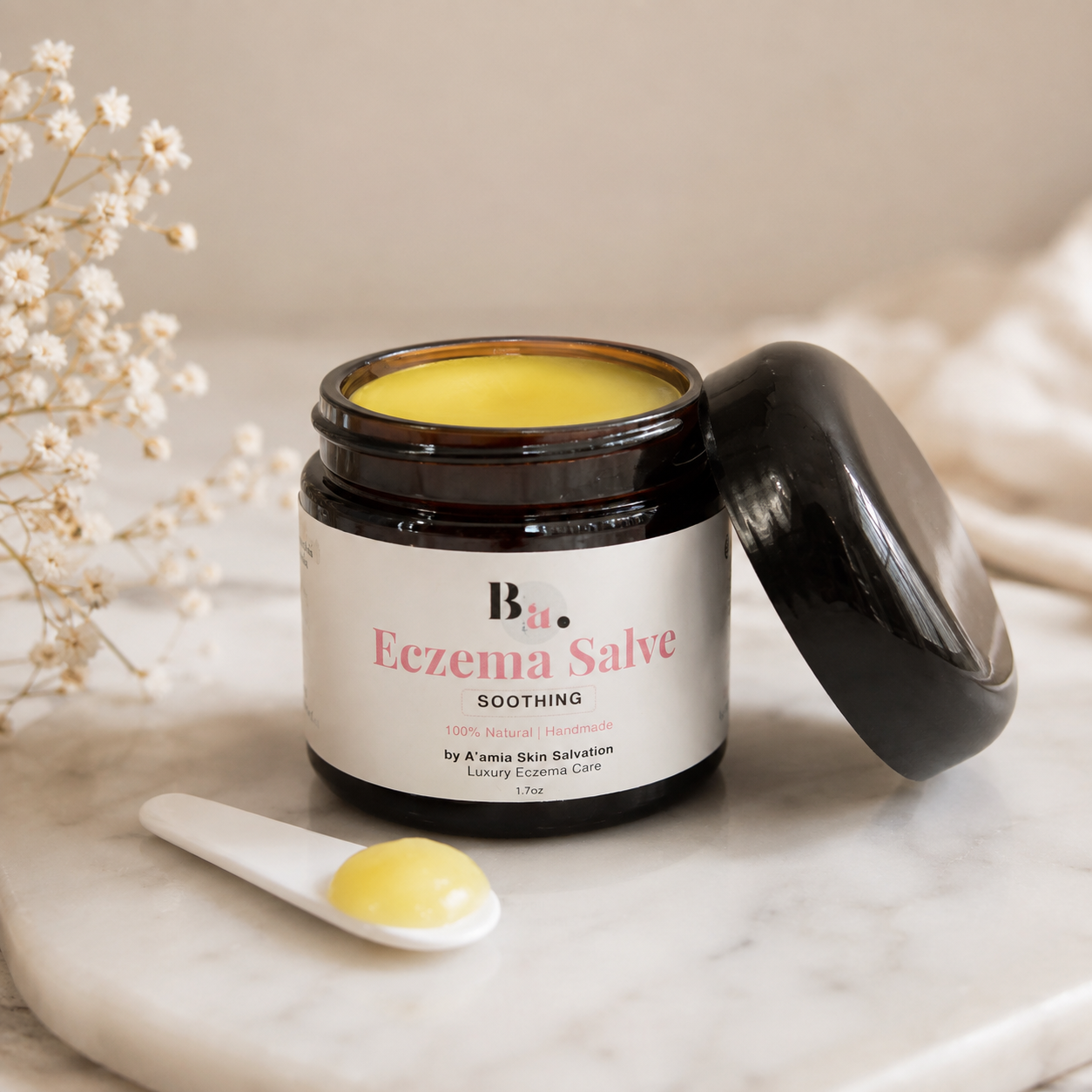 Open jar of by A’amia Eczema Salve with glossy balm texture and beauty spatula on marble.