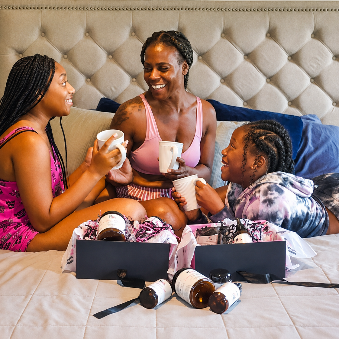 by A'amia founders sitting together on a bed with a tufted headboard, holding mugs and interacting with open gift boxes containing skincare or beauty products. The scene is indoors and styled as a lifestyle shot rather than a clean isolated product photo.